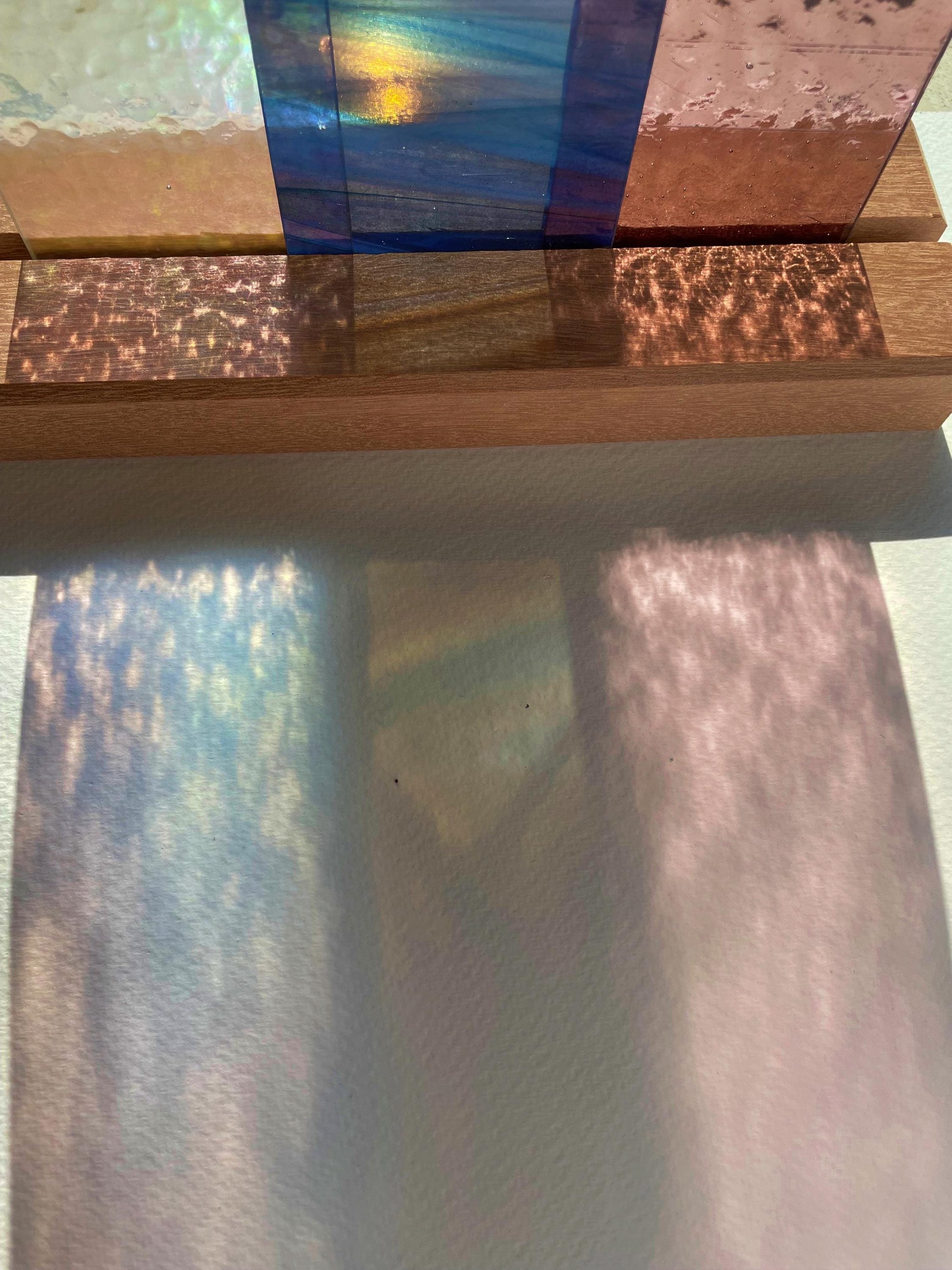 Eine Sammlung durchscheinender, farbiger Glasplatten auf einer Holzoberfläche, die bunte Schatten werfen. Die Muster ahmen die Textur und Farben des Glases nach und schaffen ein visuell ansprechendes Spiel aus Licht und Farbe